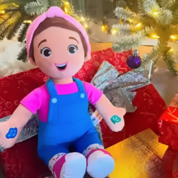 Plush toy with pink top and blue overalls sitting in front of a decorated Christmas tree.